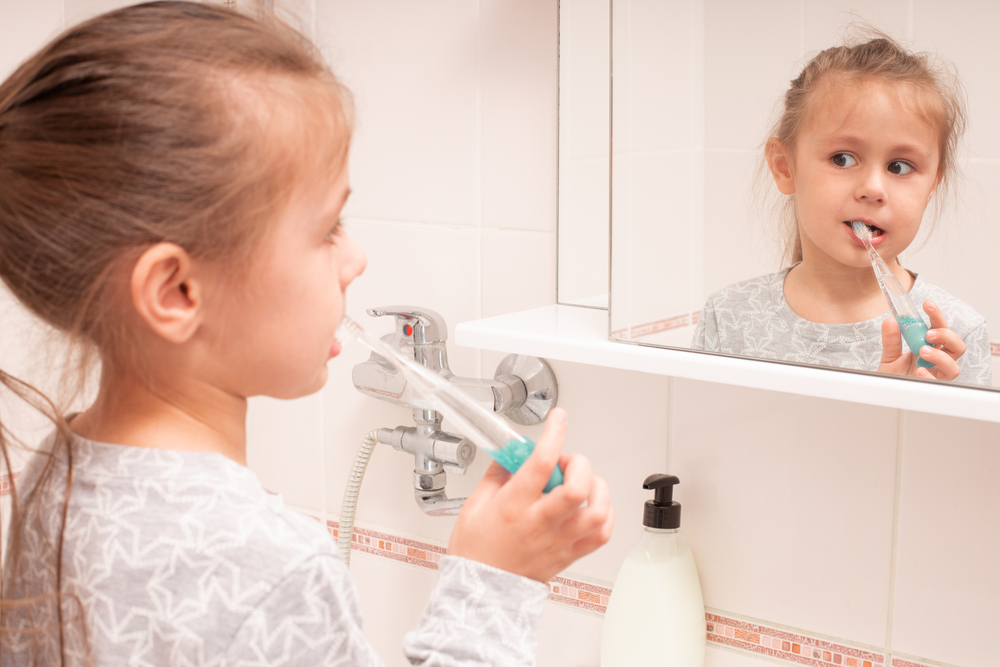  Elementary-aged girl stands at the bathroom sink and looks in mirror while brushing her teeth to stop childhood tooth decay.