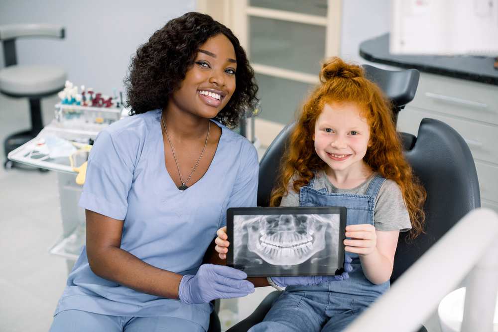  Dentist wearing scrubs and gloves sits next to a young girl in the dental chair, who holds X-ray image of girl’s healthy teeth.