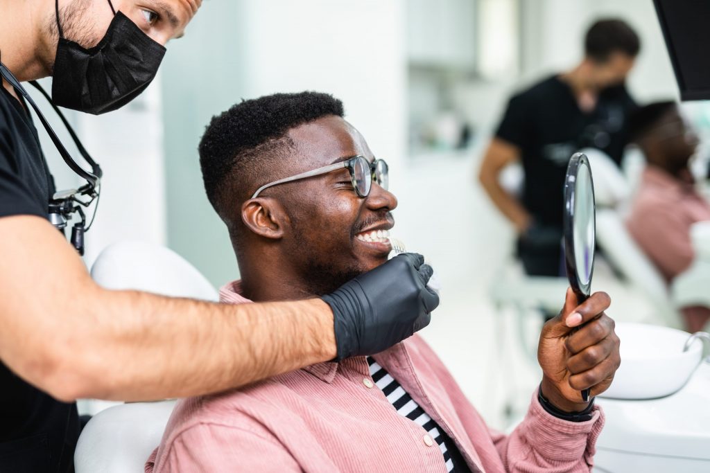 Man sits in dental chair after periodontal treatment, smiling at his reflection in hand mirror, masked periodontist behind him.