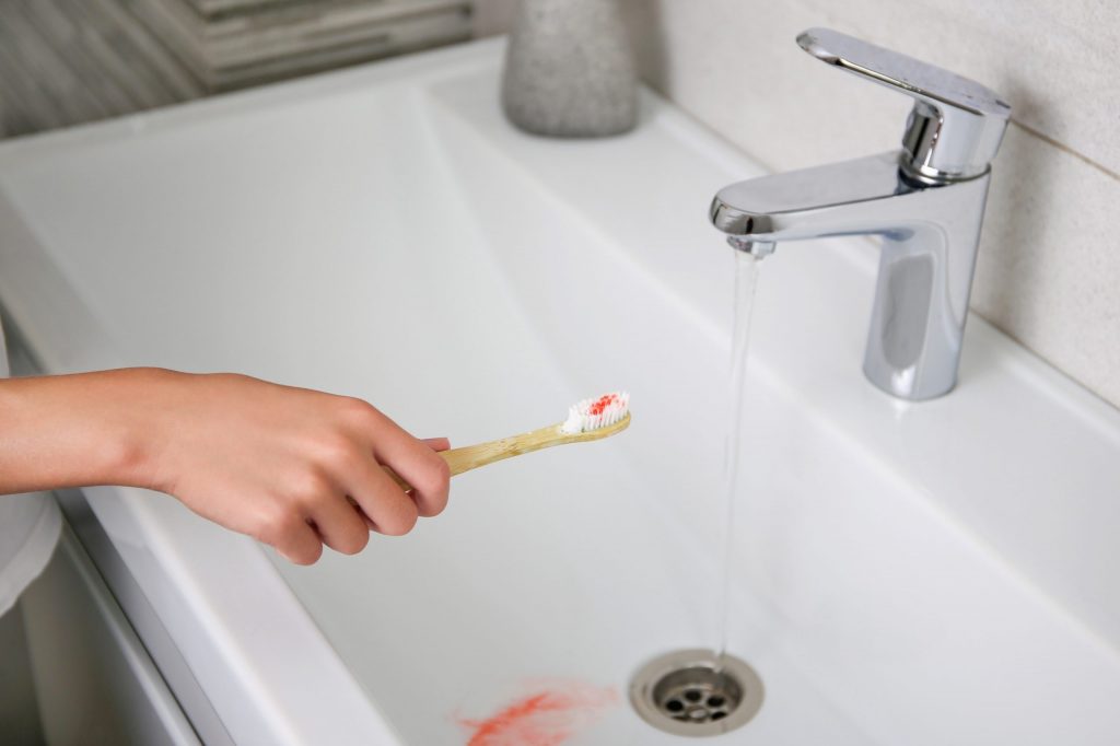 Hand holds a toothbrush with blood on it, a warning sign of periodontal disease, over a bathroom sink