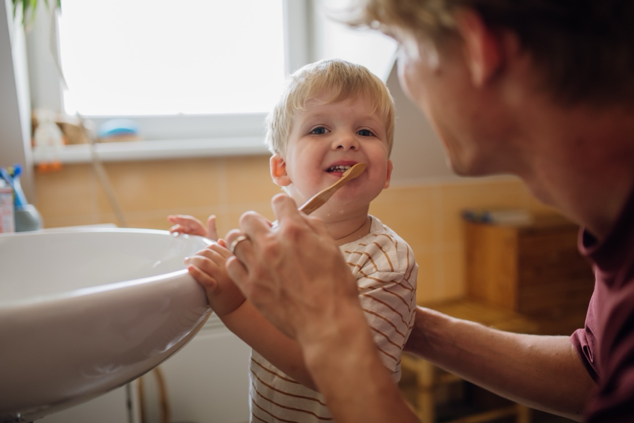 Father kneels beside toddler son at sink to teach him to brush teeth, a key in preventing tooth decay in toddlers. 