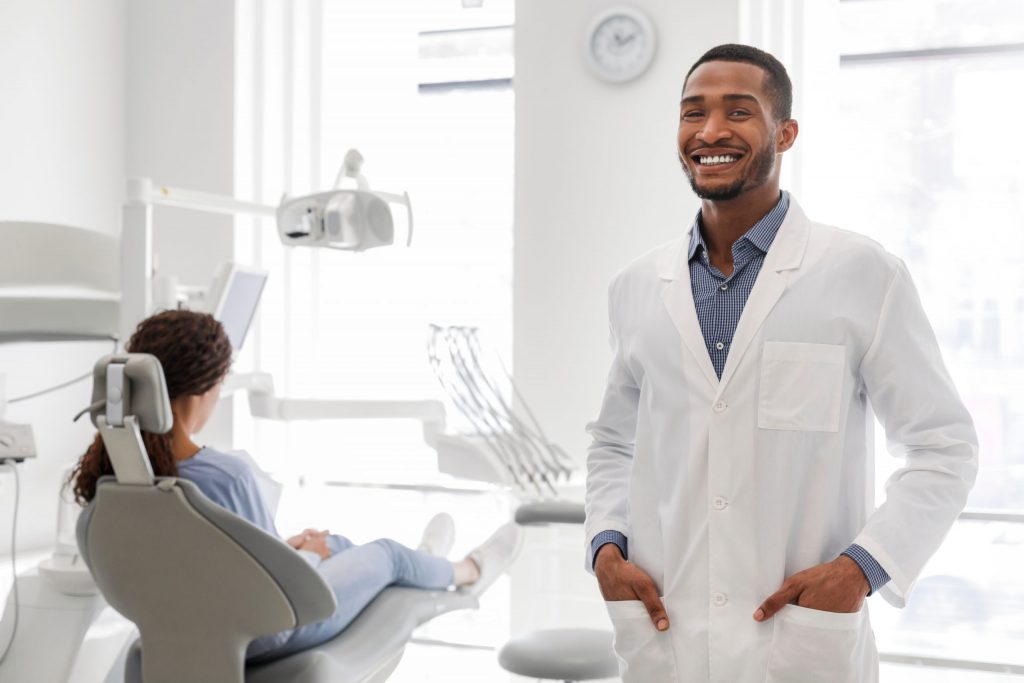  Dentist holds hand in his dental jacket pockets and smiles, woman sits in dental chair awaiting cleaning and examination.