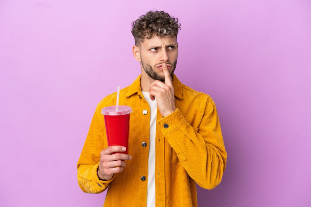  Man about to drink an extra large cup of soda holds left finger to lips as if wondering, “Is soda bad for your teeth?”