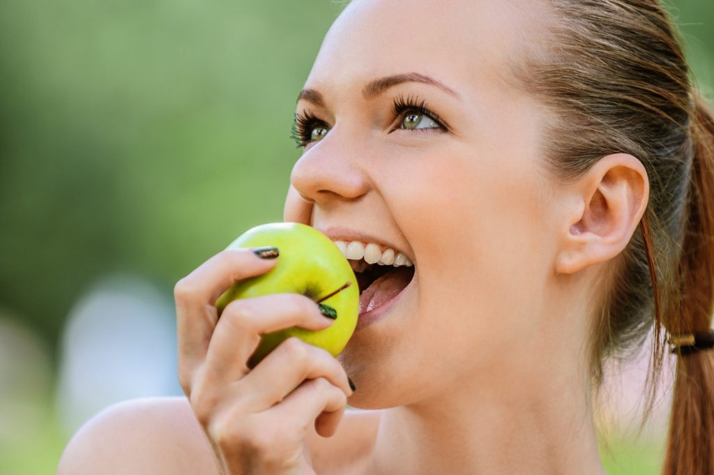  A young woman smiles as she bites into an apple because she knows it’s an enamel-strengthening food. 