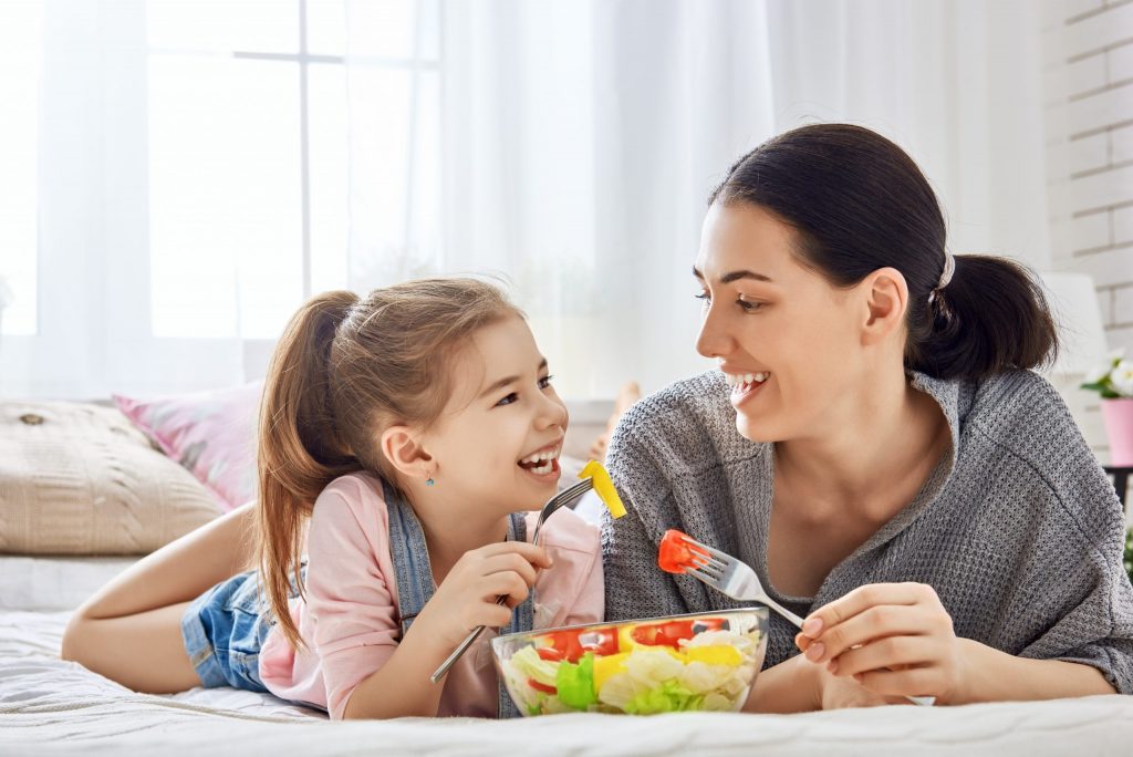 A mom and young daughter smile as they eat a salad of foods that prevent tooth decay.