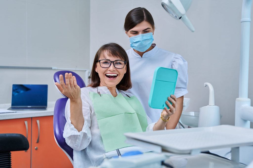 A woman in dental chair holds mirror and smiles, delighted with her whiter teeth, her teeth whitening dentist standing behind her.