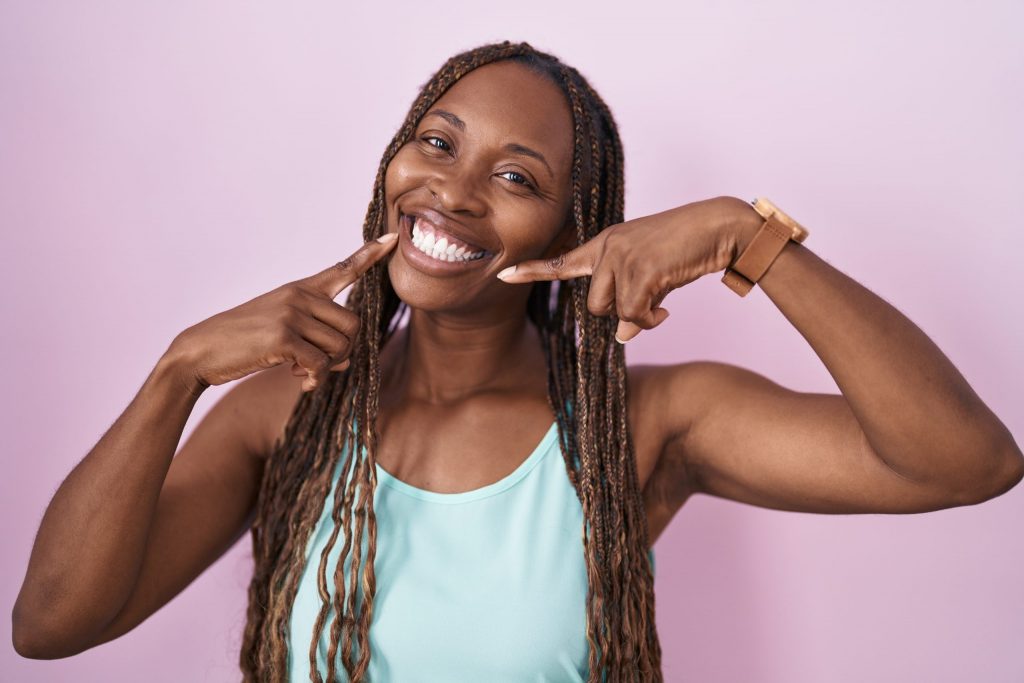 A woman points to her big, bright smile after a dentist teeth whitening treatment.   