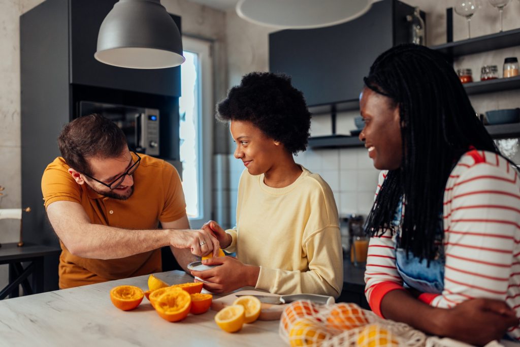 In a family’s kitchen, a mother smiles watching her husband make orange juice for their teenage daughter.