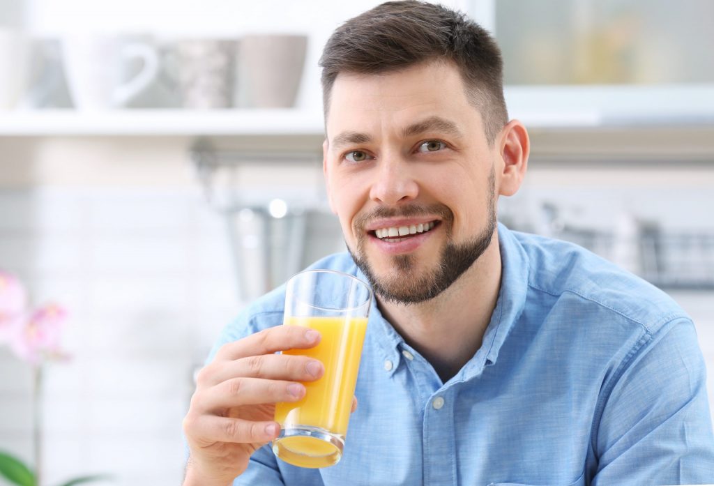  Bearded man in kitchen smiles as he drinks a glass of orange juice.
