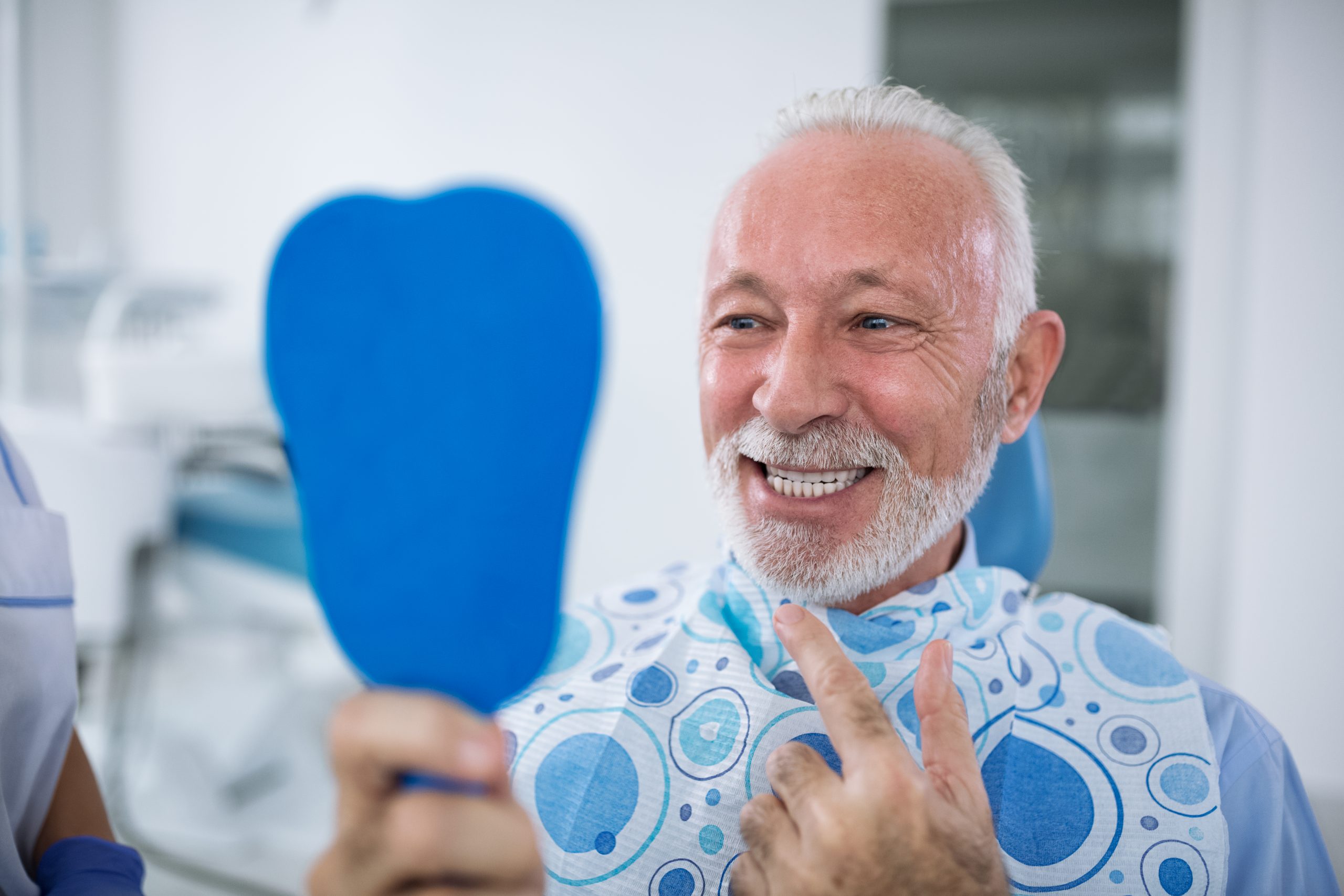A senior man smiling at the dentist looking at his teeth in the mirror.