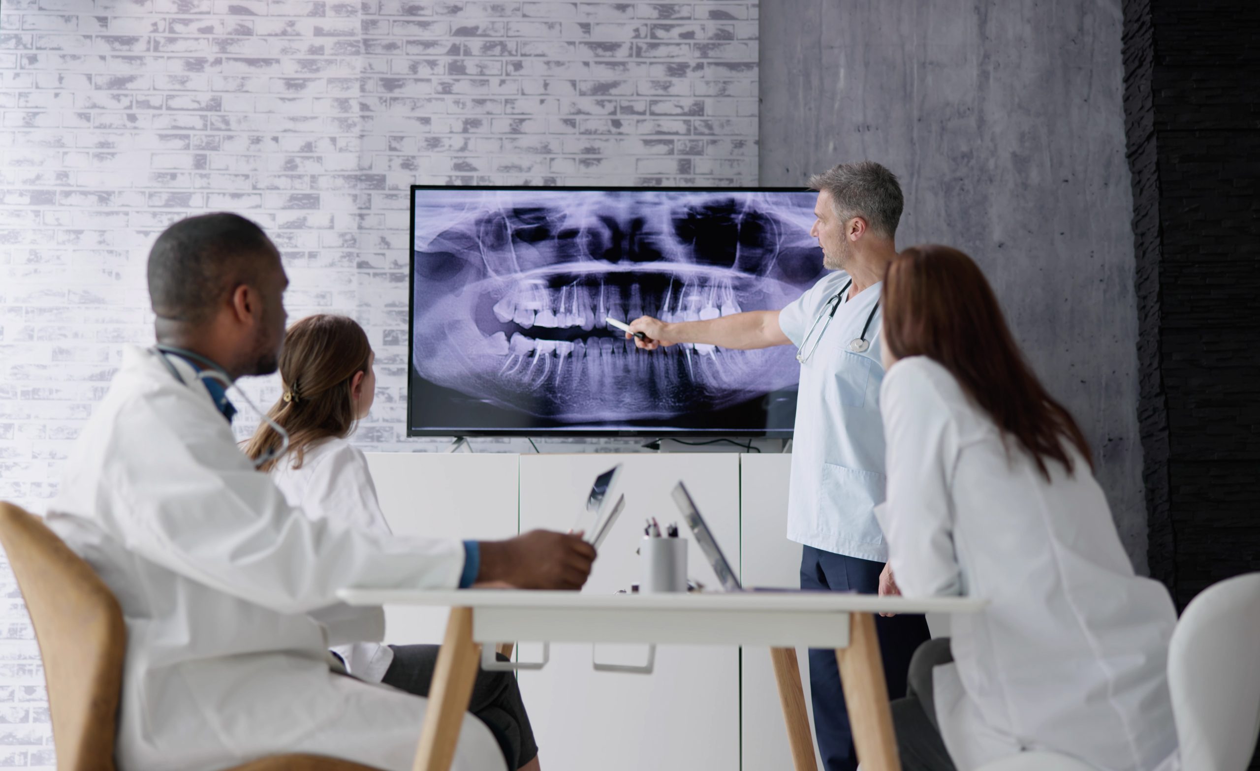 An oral medicine specialist stands in front of dental X-rays on a wall-mounted monitor, consulting with three medical colleagues.