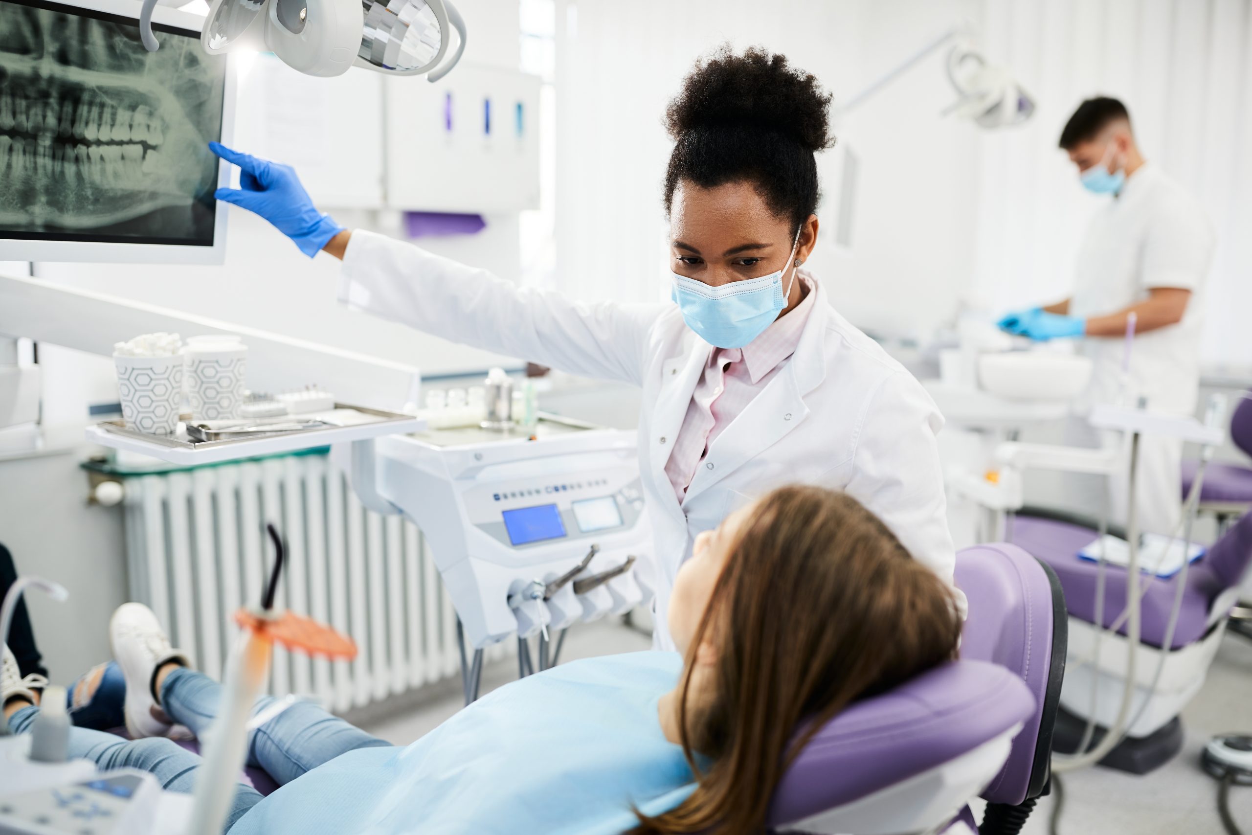 A masked and gloved Philadelphia oral medicine specialist shows dental X-rays to a girl reclining in a dental chair.