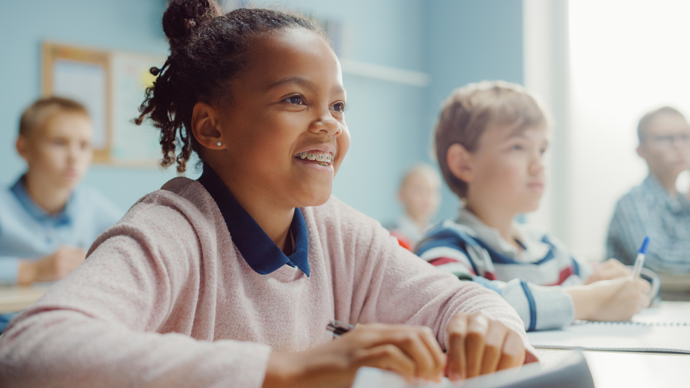 A grade-school-age girl smiles as she sits in a classroom with other students, wearing dental braces, an example of orthodontics for kids.