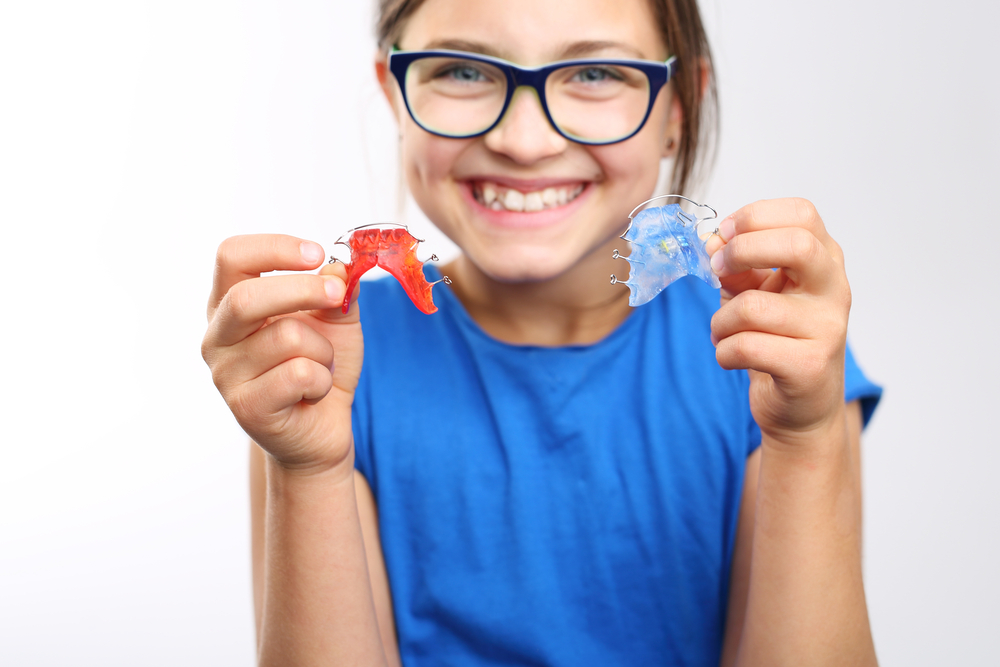 A grade-school-age girl smiles as she holds one red and one blue wire retainer, examples of orthodontics for kids.