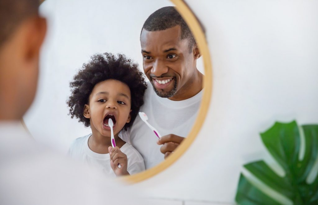 A young father and his daughter brush their teeth in a mirror.