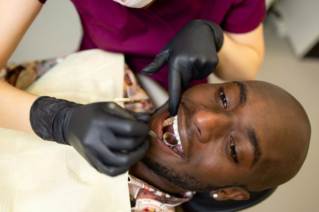 A man reclining in the dental chair in a dentist’s office is having his teeth inspected by a gloved dentist who uses a dental mirror.