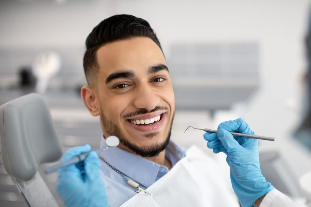 A young man smiles as a dental hygienist holds dental instruments near his mouth. 