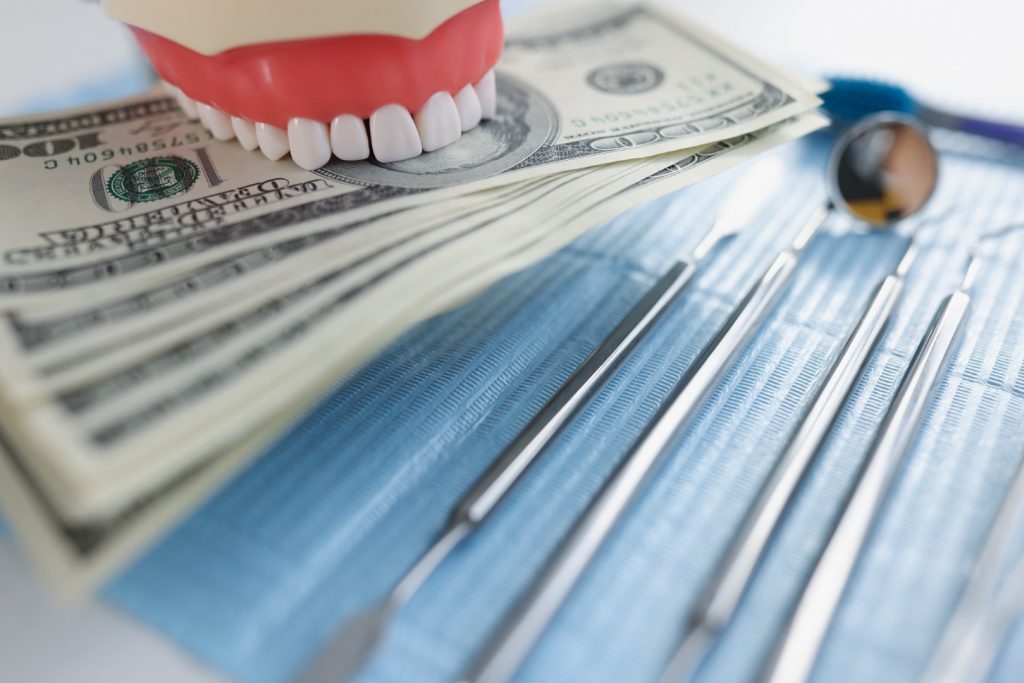 A set of teeth holding $100 bills next to dental tools.