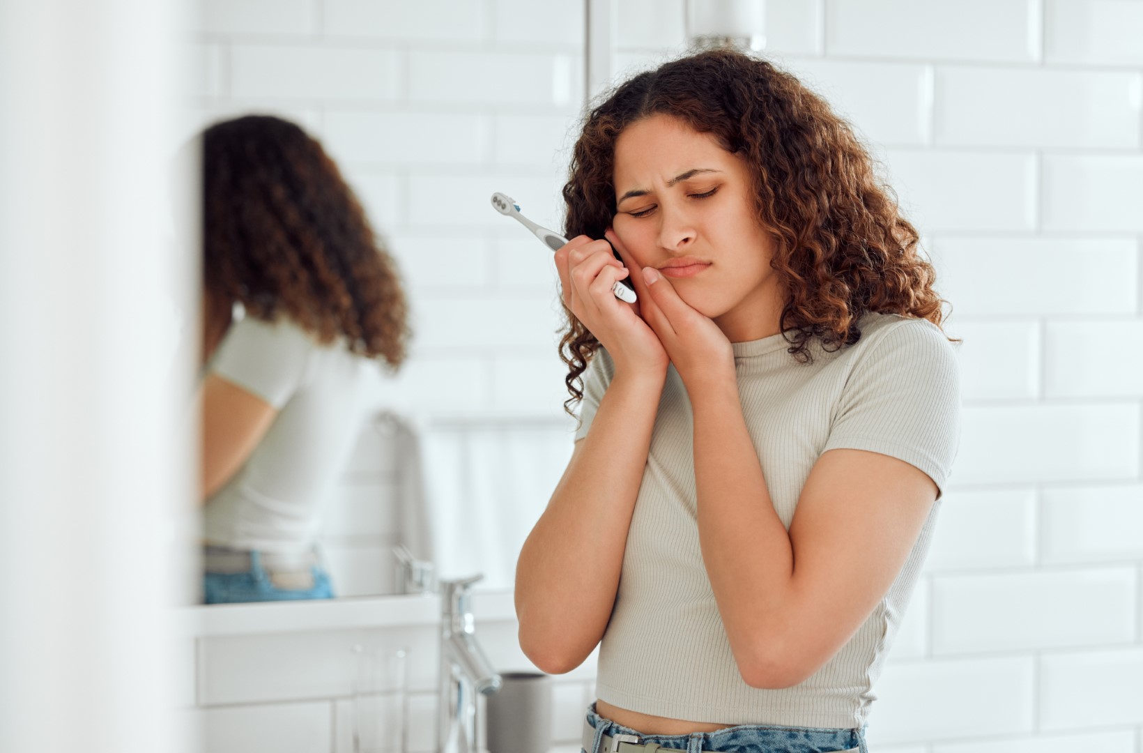 Woman holding toothbrush stops brushing her teeth and presses left hand to cheek.