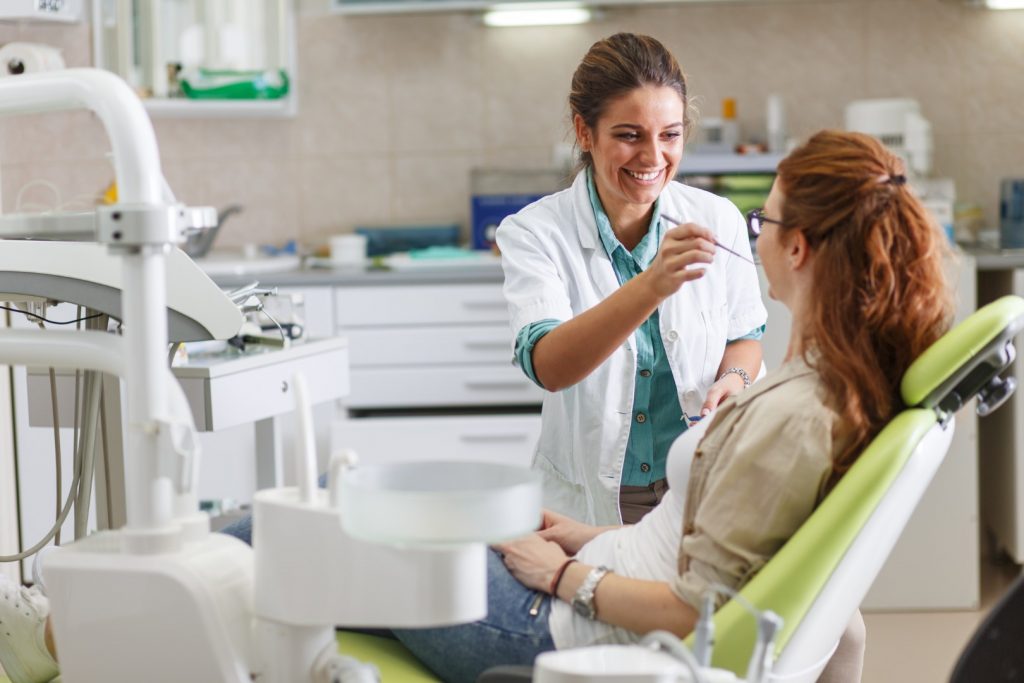 A dentist smiles as she prepares to examine mouth of patient reclining in a dental chair.