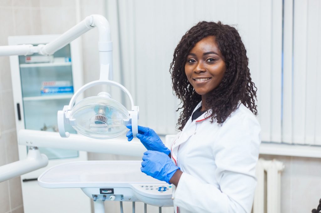 Dentist who treats TMJ disorder stands next to dental light above dental chair in her examining room.