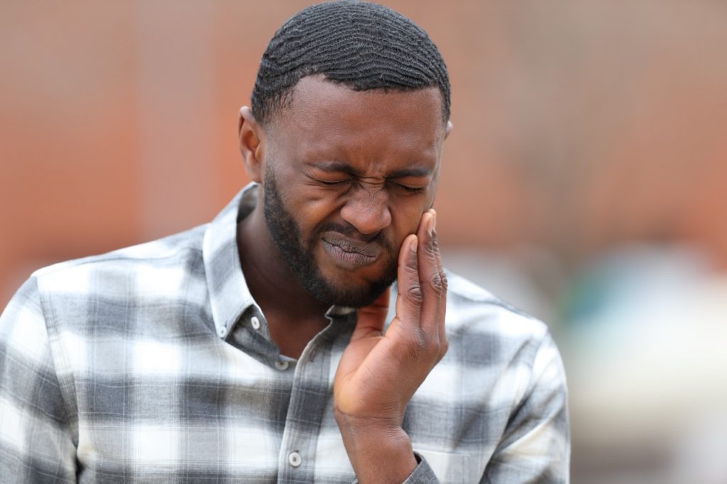 Man winces and holds his left hand to his jaw, feeling pain from TMJ disorder.