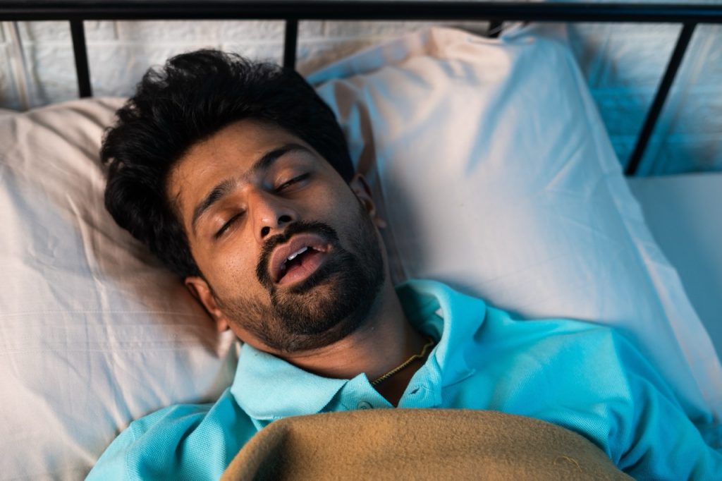 A young man lays on his back in bed, snoring loudly due to sleep apnea and dental health. 