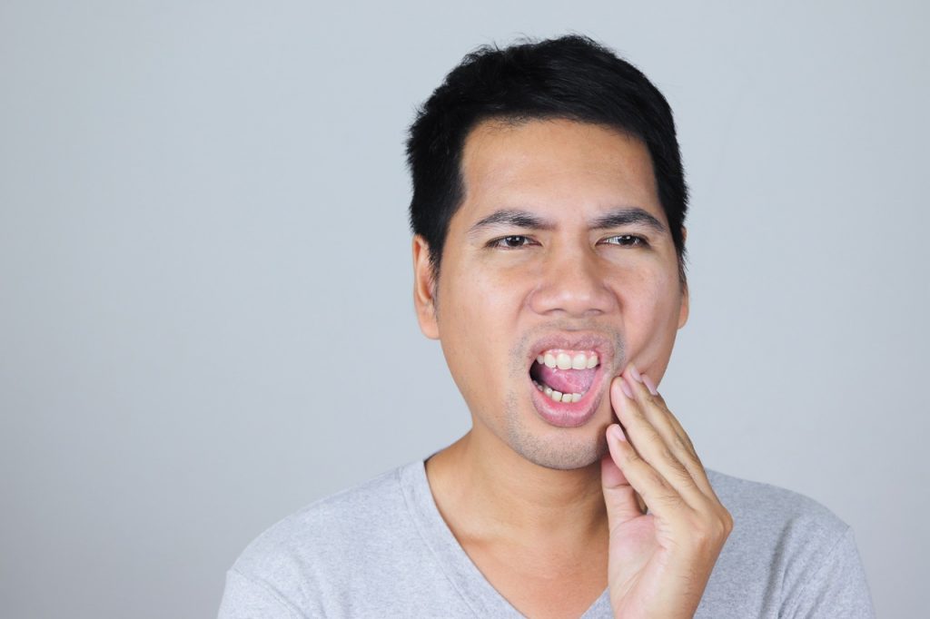 A young man holds his hand to his sore jaw from grinding teeth, which is also causing a migraine headache.