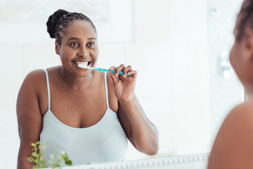 Woman stands at her bathroom mirror, smiling as she brushes her teeth to clean them rather than doing oil pulling for teeth.