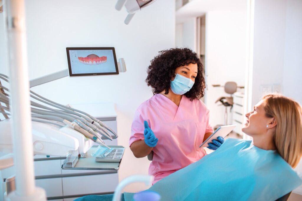 Masked oral surgeon talks to woman reclining in dental chair before her wisdom teeth removal procedure.