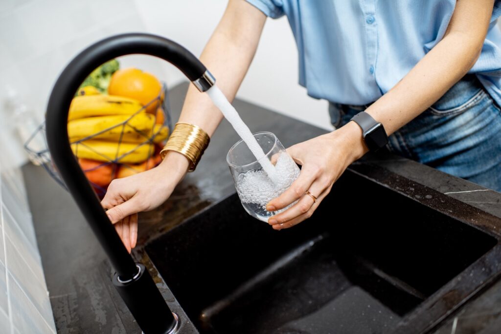 Woman stands at her kitchen sink and fills a drinking glass with fluoridated water from her tap. 