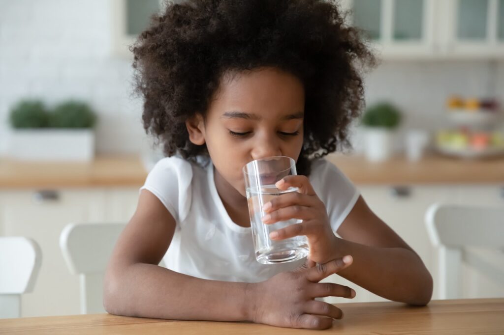 Preschool-age girl sits at kitchen table drinking glass of fluoridated water. 