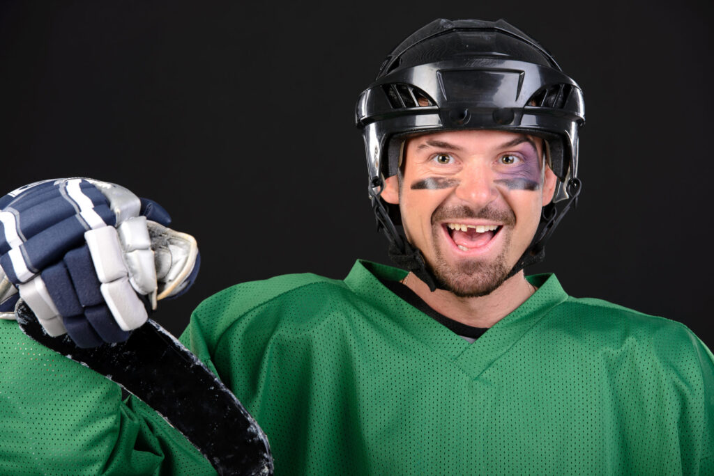 Hockey player in jersey, helmet, and gloves holds goalie stick, smiles showing where a front tooth was knocked out.