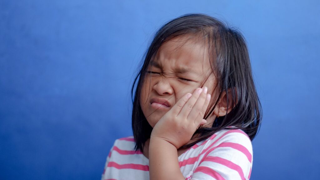 Girl winces as she presses her left hand to her cheek, indicating distress from severe toothache pain, a dental emergency.