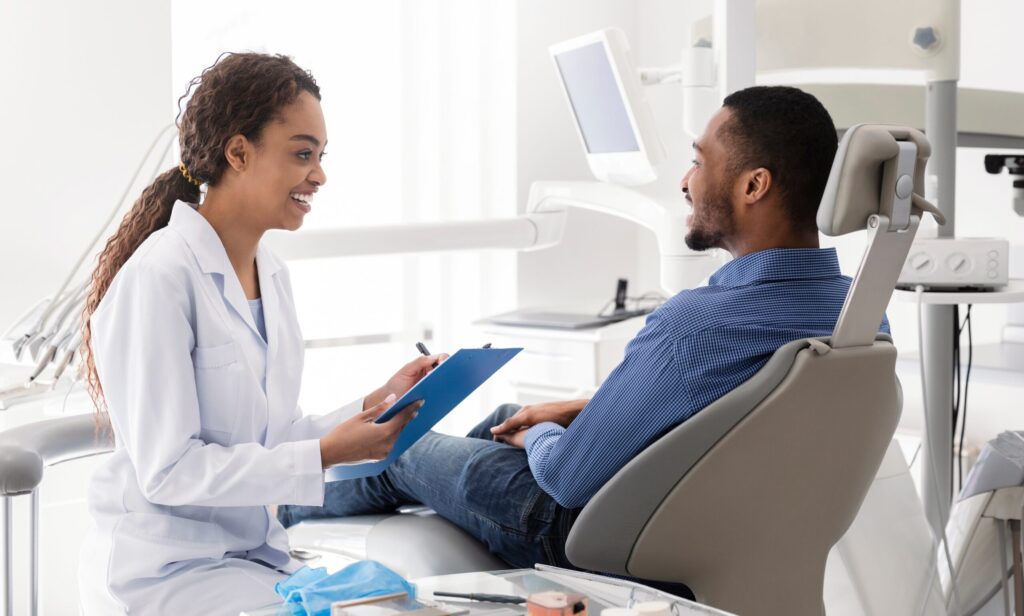 Dentist holding clipboard talks with man reclining in dental chair about his need for emergency dental care.