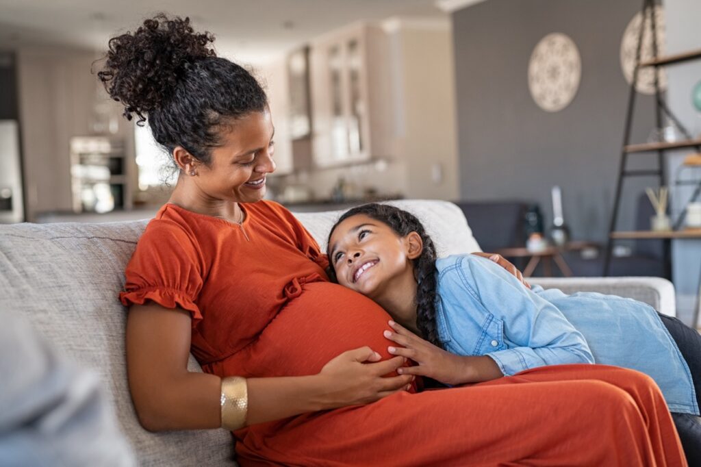 Pregnant woman and her daughter sit on couch and smile, the daughter hugging her mother’s belly. 