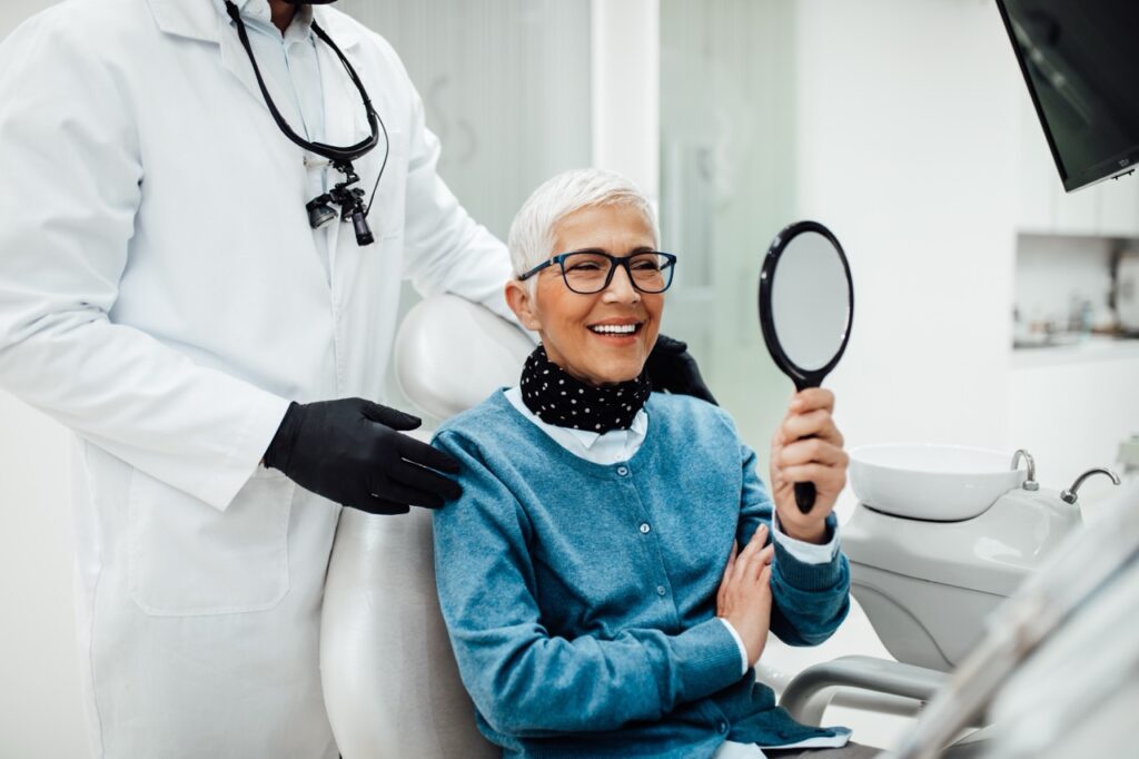 Postmenopausal woman sits in a dental chair and holds a hand mirror, smiling at how her teeth look bright and healthy.