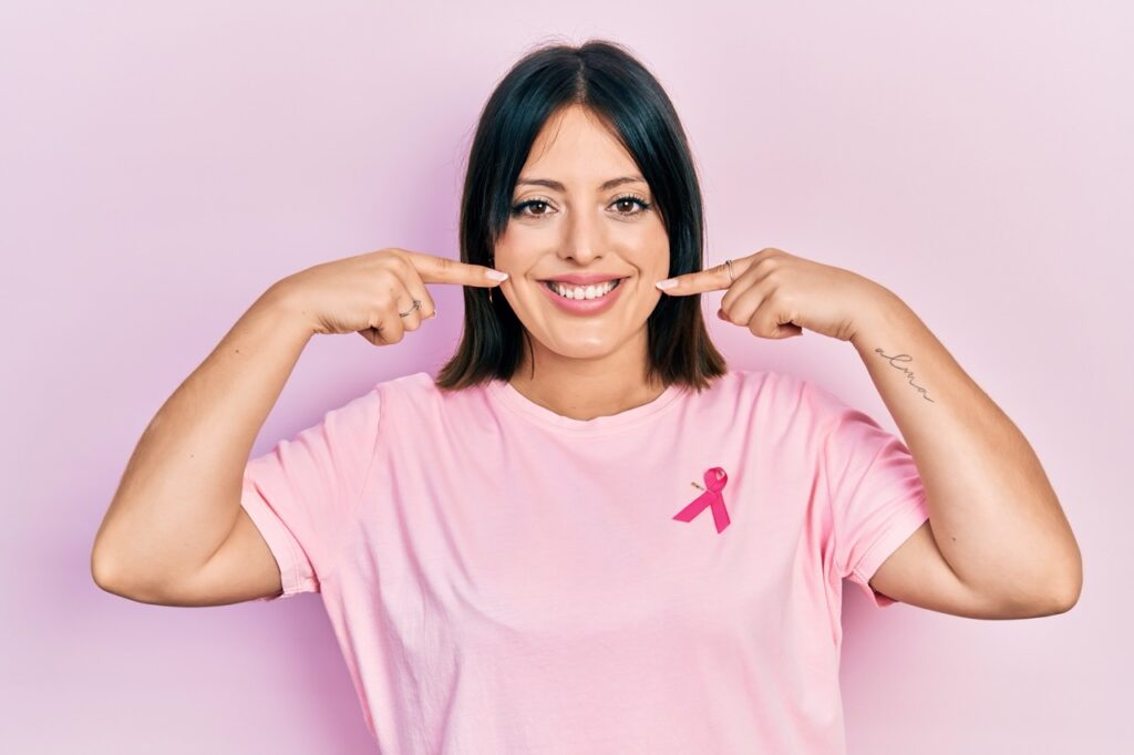 Breast cancer patient wearing pink T-shirt and ribbon points with both hands to her bright and healthy smile.