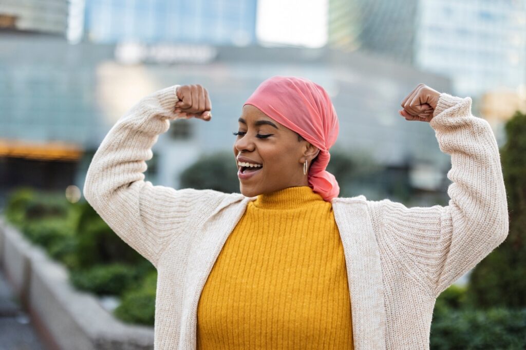 Breast cancer patient wearing scarf on her head smiles and lifts her arm in triumph after successful treatment.