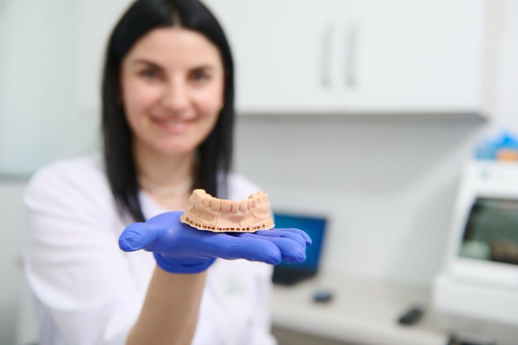 Prosthodontist smiles as she holds a model of lower set of teeth in her hand, used for fabricating dental prosthetics.