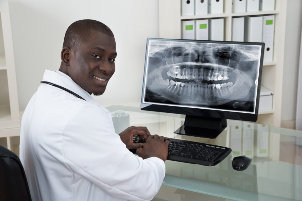 Prosthodontist sits at desktop computer, viewing X-ray of upper and lower jaws as he designs dental prosthetics. 