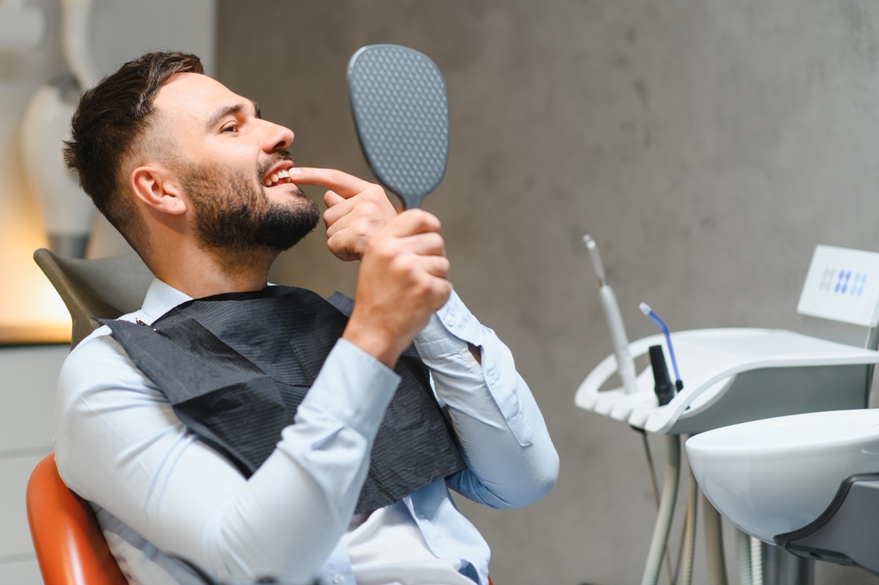 Man sitting in dental chair uses hand mirror to admire restoration of a dental crown.
