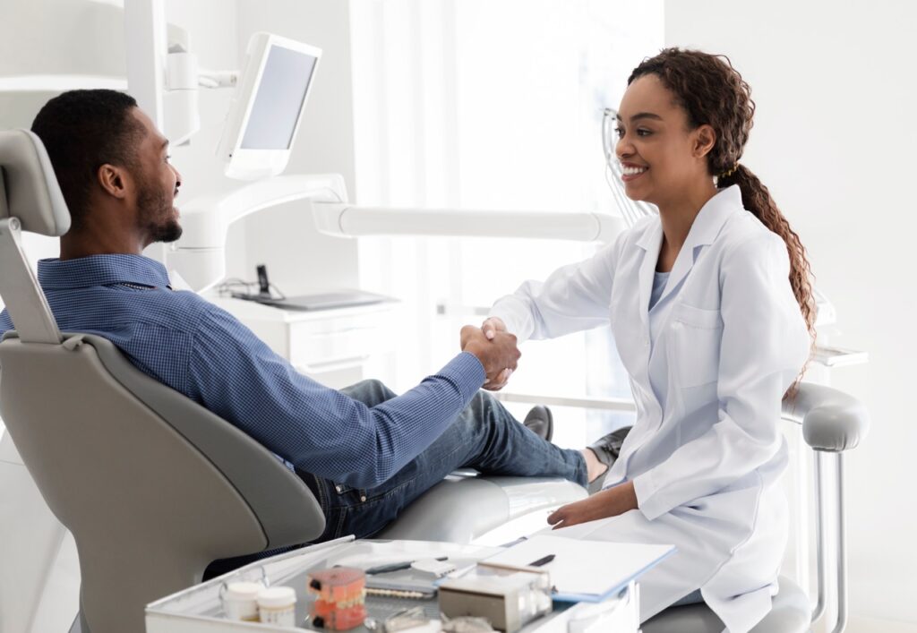 Man reclines in dental chair and shakes the hand of his dentist after she has cleaned and examined his teeth.