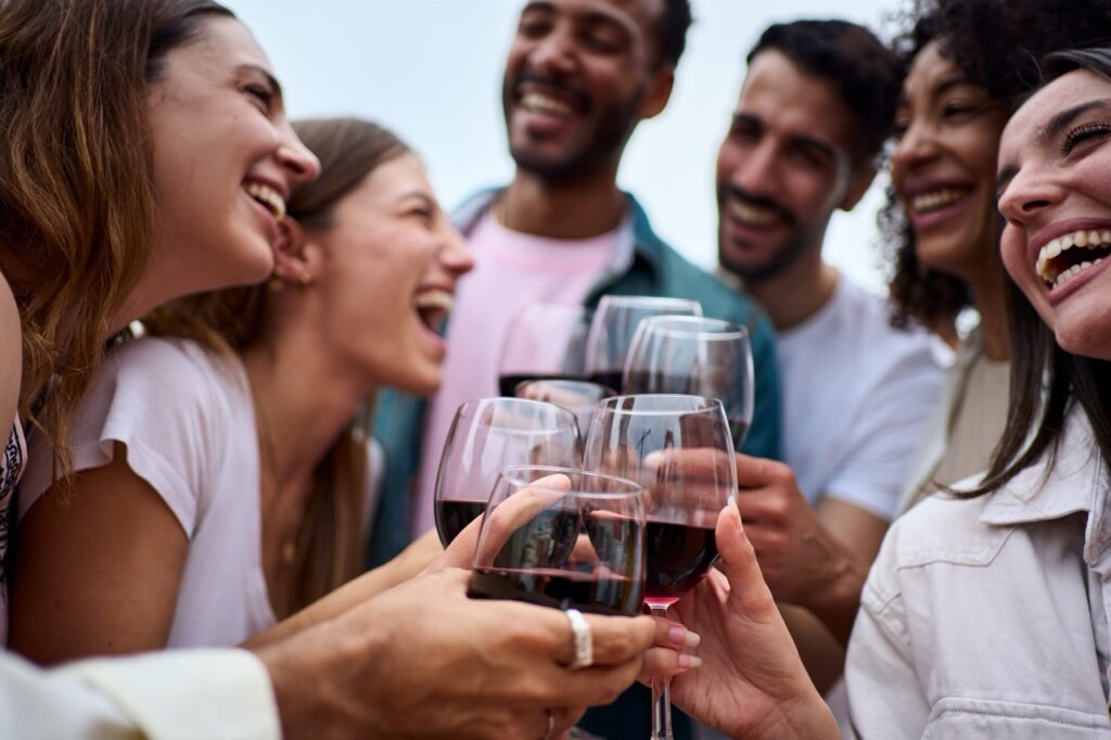 Six friends in semicircle laugh and toast with glasses of red wine. Staining is one of the effects of alcohol on teeth. 