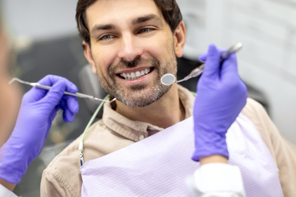 Man reclines in dental chair and smiles as gloved dentist holds dental pick and mirror, preparing to examine his teeth.