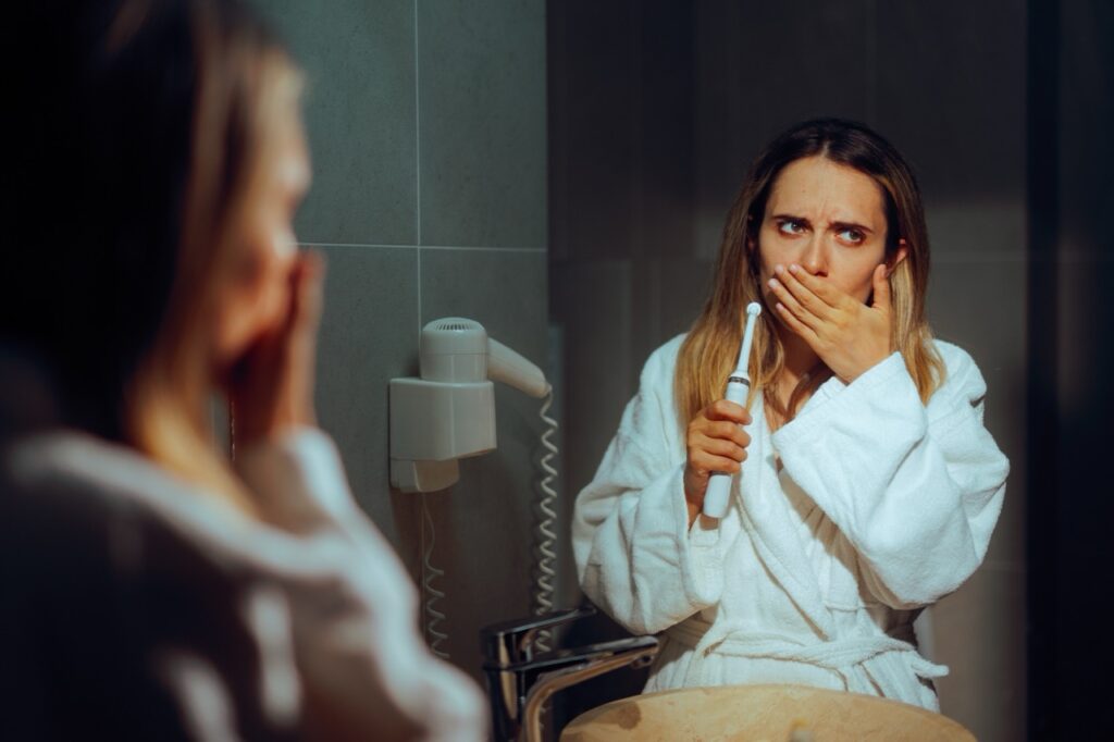 Woman holds electric toothbrush in one hand and the other hand to her mouth because she feels pain when brushing. 