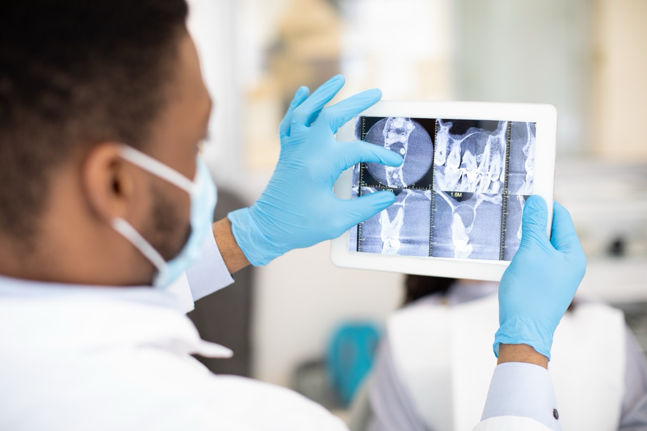 Gloved and masked dentist examines digital radiography images of patient’s teeth on his tablet computer.