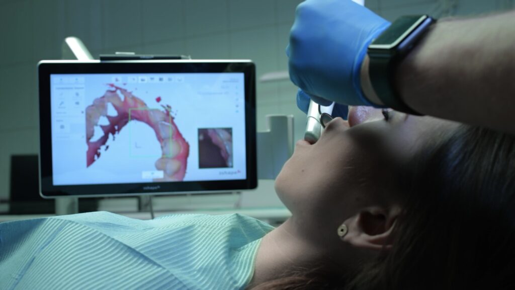 Dentist uses intraoral scanners to create digital impression of teeth of woman reclining in dental chair.
