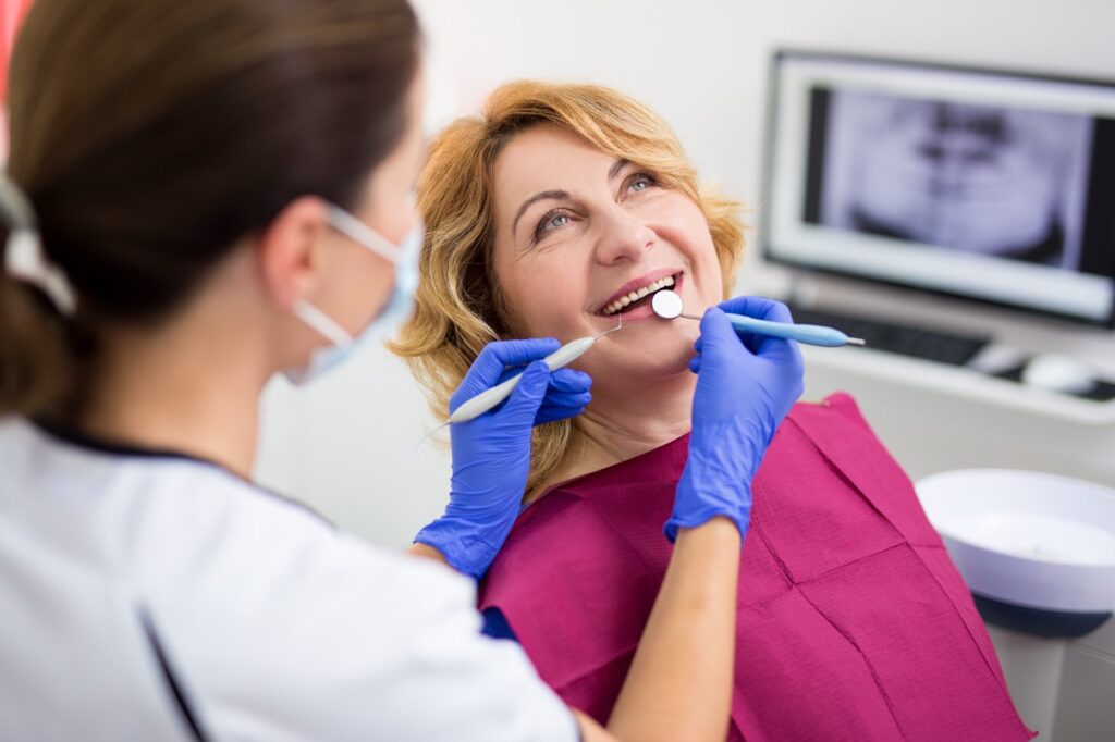 Woman reclines in dental chair as dentist examines her teeth, AI-generated X-rays of teeth on monitor behind. 