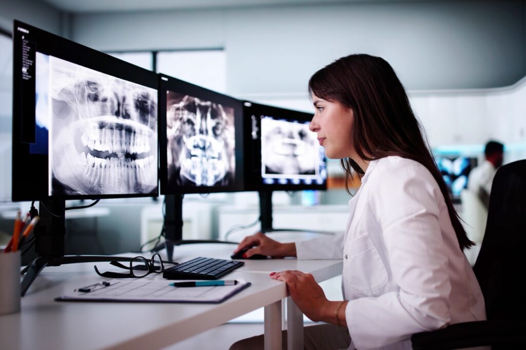 Assisted by dental artificial intelligence, dentist examines her patient’s X-rays on computer monitors.